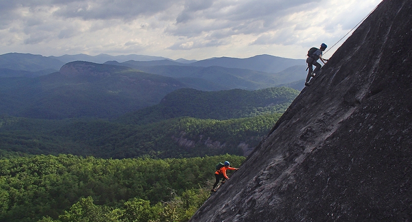 Two people wearing safety gear are secured by ropes as they make their way up a rock incline. Behind them is a vast mountainous area covered in thick green trees. 